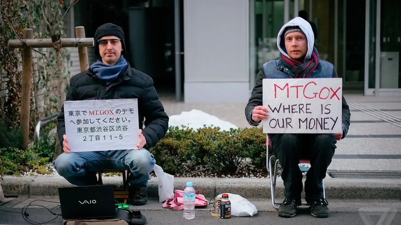 Image of two men sitting near the sidewalk seemingly protesting holding signs about Mt Gox with a laptop on the ground. One sign says 'Mt Gox Where is our money', while the other mentions Mt Gox within a seemingly Chinese text. 