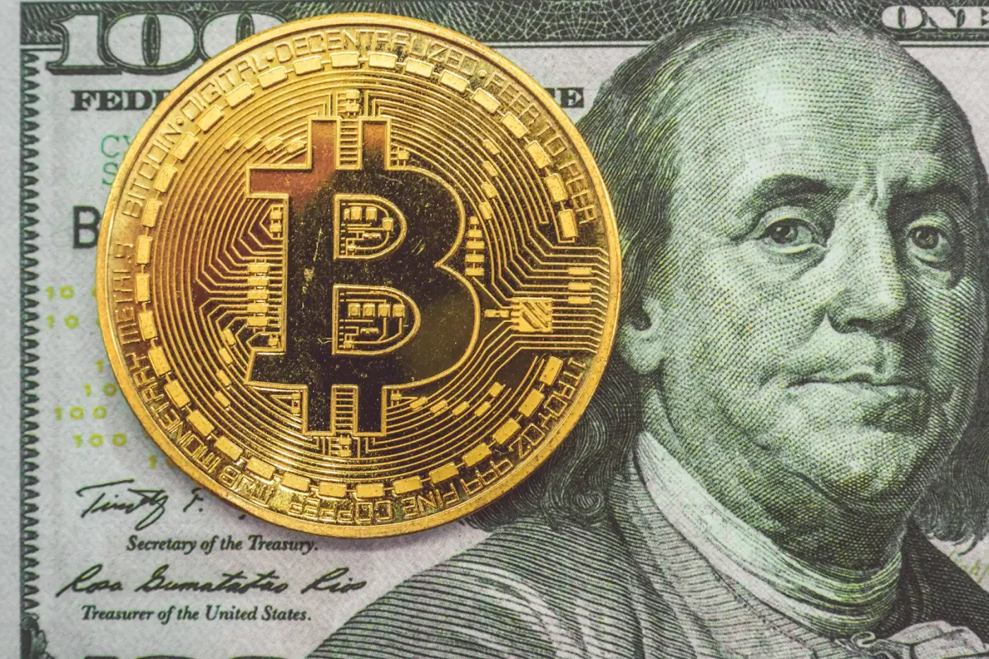Image of a Bitcoin's symbol on a gold coin placed on top of a US $100 bill